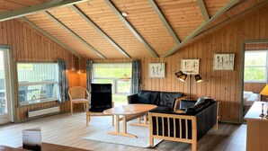 A cozy room with a wooden ceiling, a black leather sofa, a wooden table, and a chair. There are windows with curtains, a lamp, and framed pictures on the wall. - "Juha" -  from the sea by Interhome (Hvide Sande)