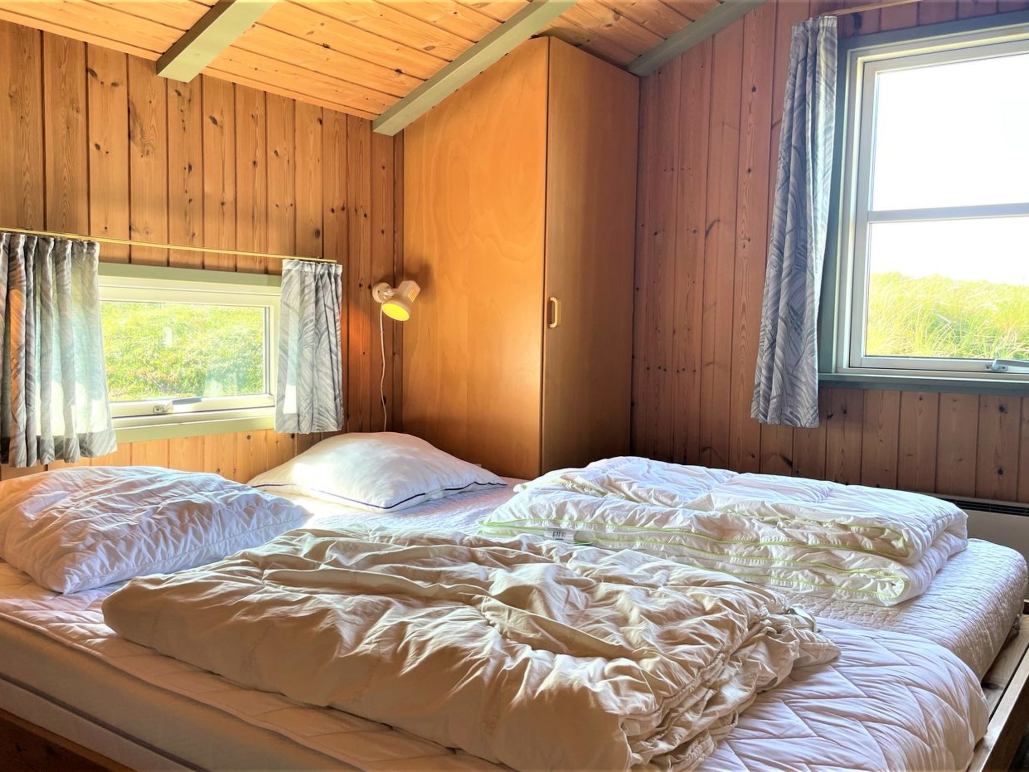 Quarto de cabana de madeira com duas camas, uma janela e um abajur fixado na parede.