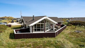 Une maison moderne dotée d’une grande terrasse, entourée d’une clôture en bois, et une allée en gravier menant à l’entrée.