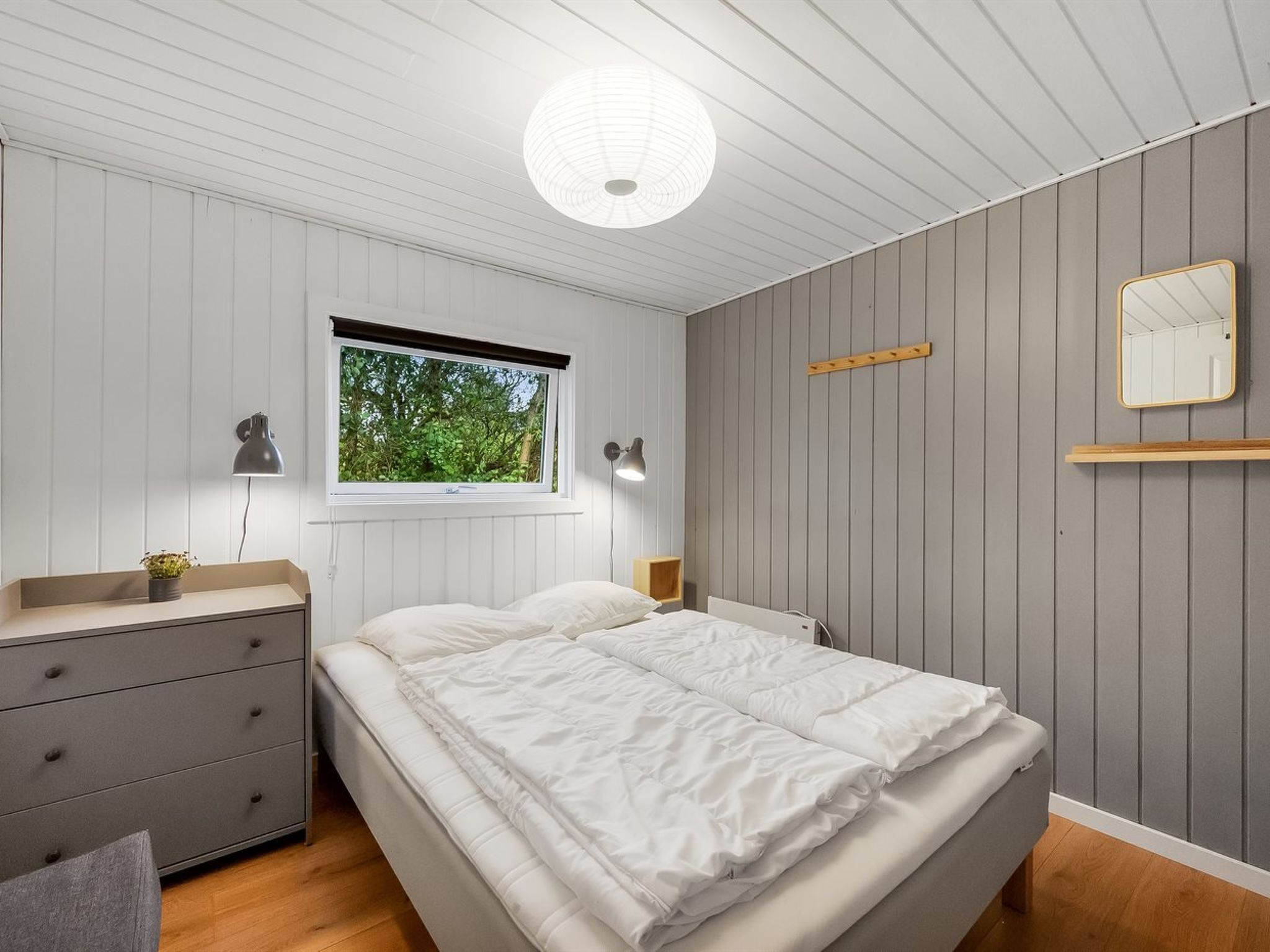 Un dormitorio con cama, lámparas de noche, ventana, mesita de noche, cómoda y espejo en la pared.