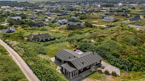 Vista aérea de una zona residencial con casas, una casa moderna destacada con un amplio balcón, y un camino de tierra.