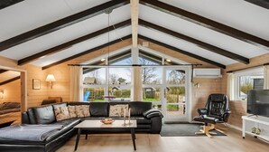 Una sala de estar acogedora con un sofá de cuero negro, una mesa de centro y un televisor. Se ve una ventana grande con vistas al exterior.