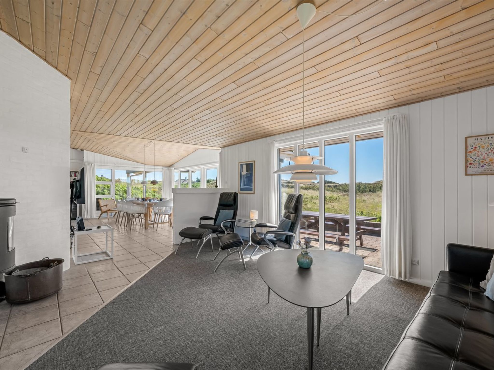 A spacious room with a wooden ceiling, a dining area with chairs and a table, a sofa, and a large window offering a view of the outdoors.