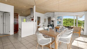 Un vaste espace de salle à manger avec une table en bois et des chaises blanches, une cuisine en arrière-plan et de grandes fenêtres donnant sur un paysage verdoyant.