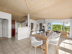 A spacious dining area with a wooden table and white chairs, a kitchen in the background, and large windows offering a view of greenery.