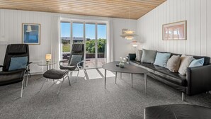 Une pièce avec un plafond en bois, une grande fenêtre, un canapé en cuir noir, une table ronde et deux chaises.