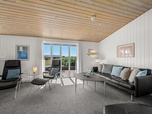 A room with a wooden ceiling, a large window, a black leather sofa, a round table, and two chairs.