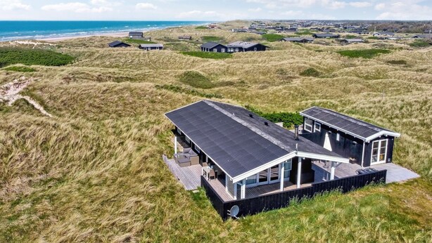 A modern house with a sloped roof, large windows, and a deck, surrounded by grassy dunes and a beach in the background.