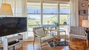 A cozy living room with a large flat-screen TV, wicker chairs, a round table, and a view of the outdoors through large windows.