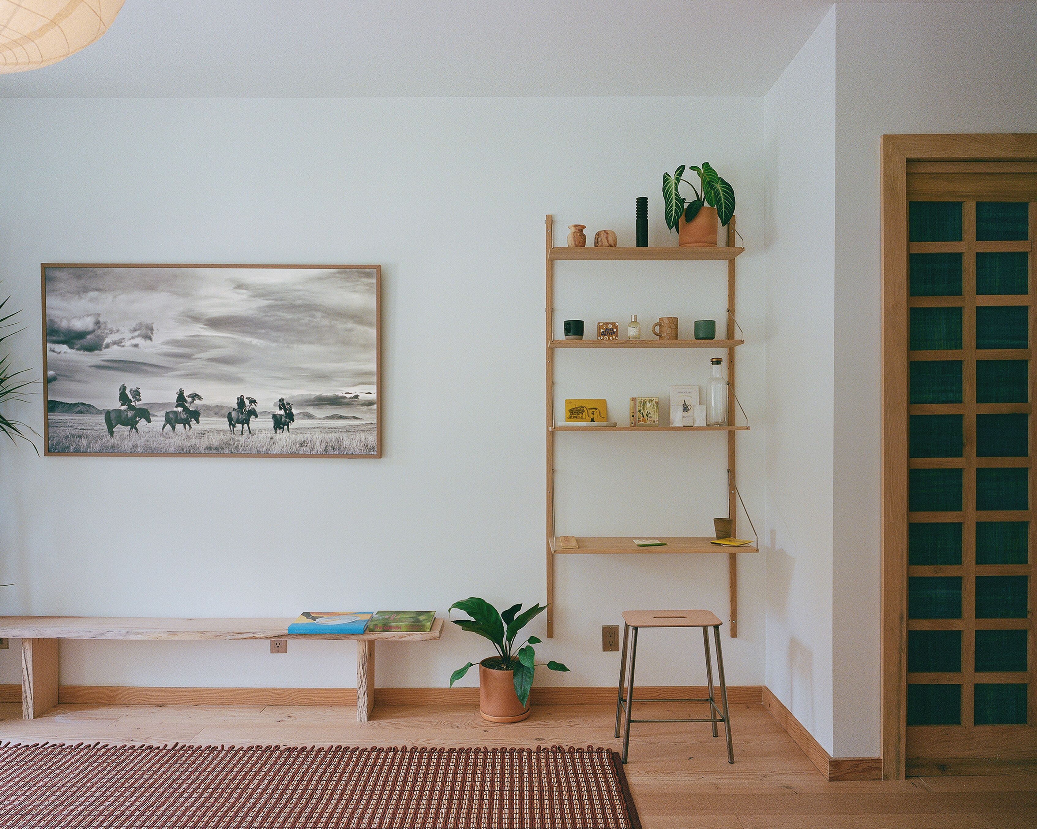 Ein Zimmer mit einer hölzernen Bank, einem Blumentopf, einem Hocker und einem Regal mit diversen Gegenständen.
