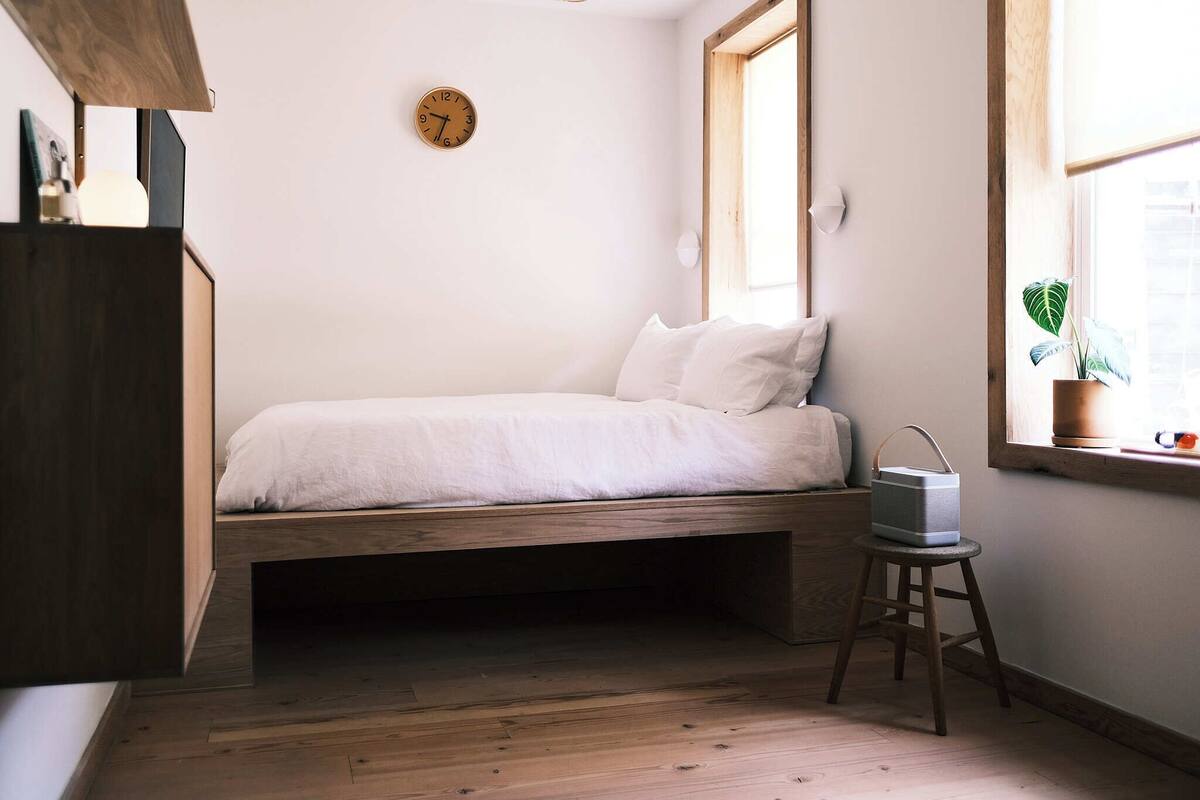 A small, minimalist bedroom with a wooden bed frame, a bedside table, a clock on the wall, and a window with a plant.