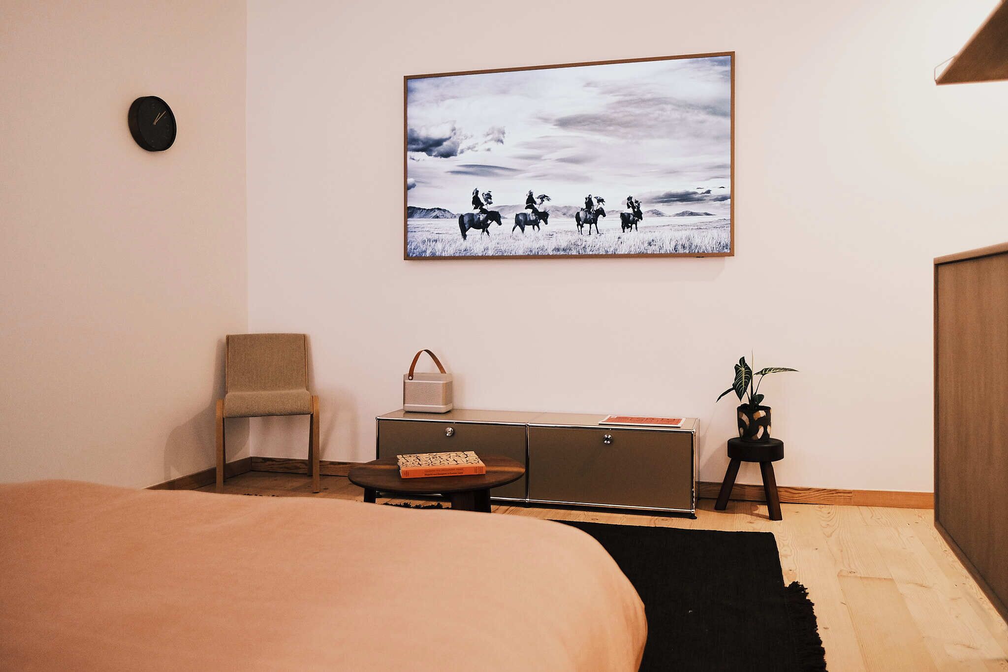 A bedroom with a bed, a chair, a small table with a book and a bag, a dresser, a plant, and a painting of horse riders on the wall.