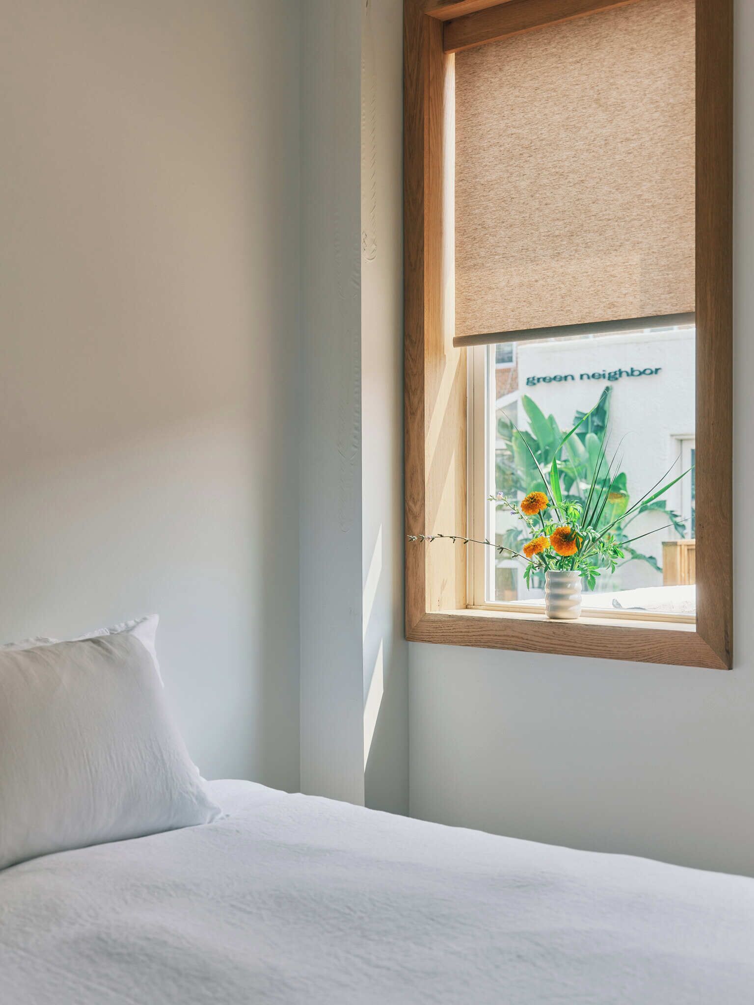 A bedroom with a bed, a window with a view of a building labeled "green neighbor," and a vase with flowers on the windowsill.