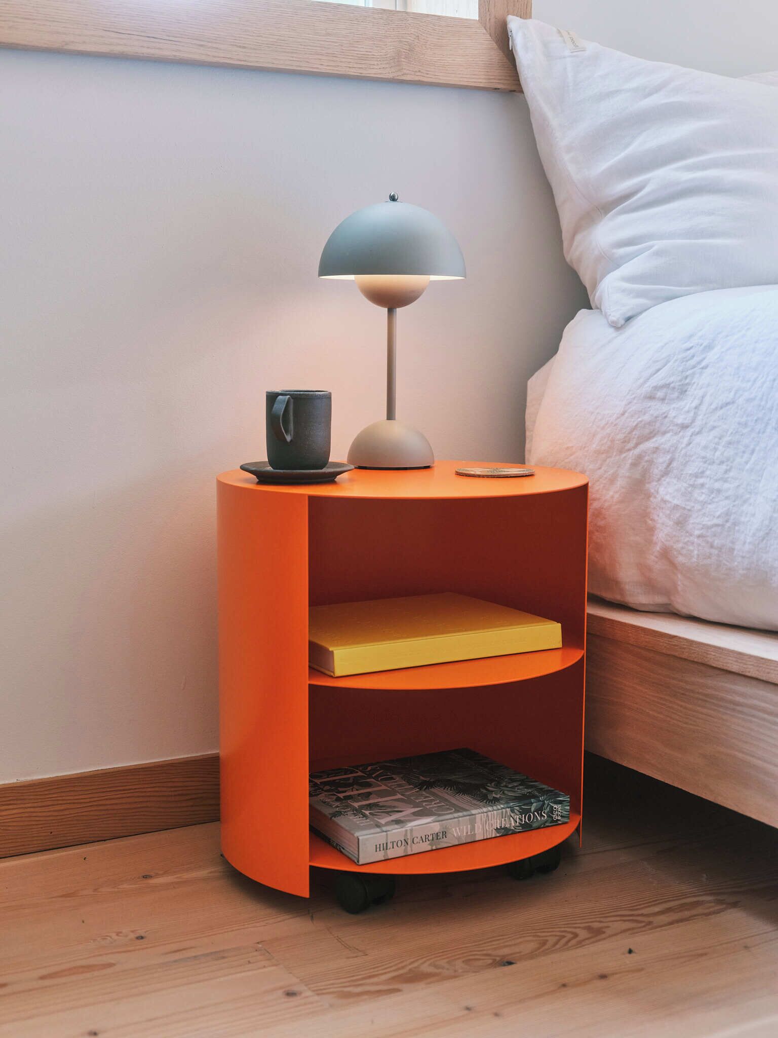 A modern bedroom with a wooden headboard, a bed with white bedding, an orange nightstand with a lamp, a mug, and books.
