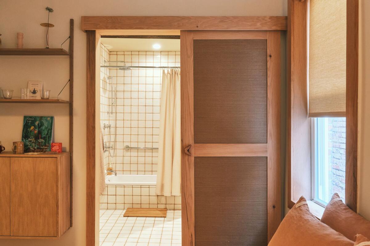 A bedroom with a wooden wardrobe, a bedside table with a book, a window with a view, and a bathroom with a shower and sink.