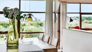 Uma sala de jantar moderna com uma mesa e cadeiras, um vaso com flores e janelas amplas com vista para o verde e uma casa.