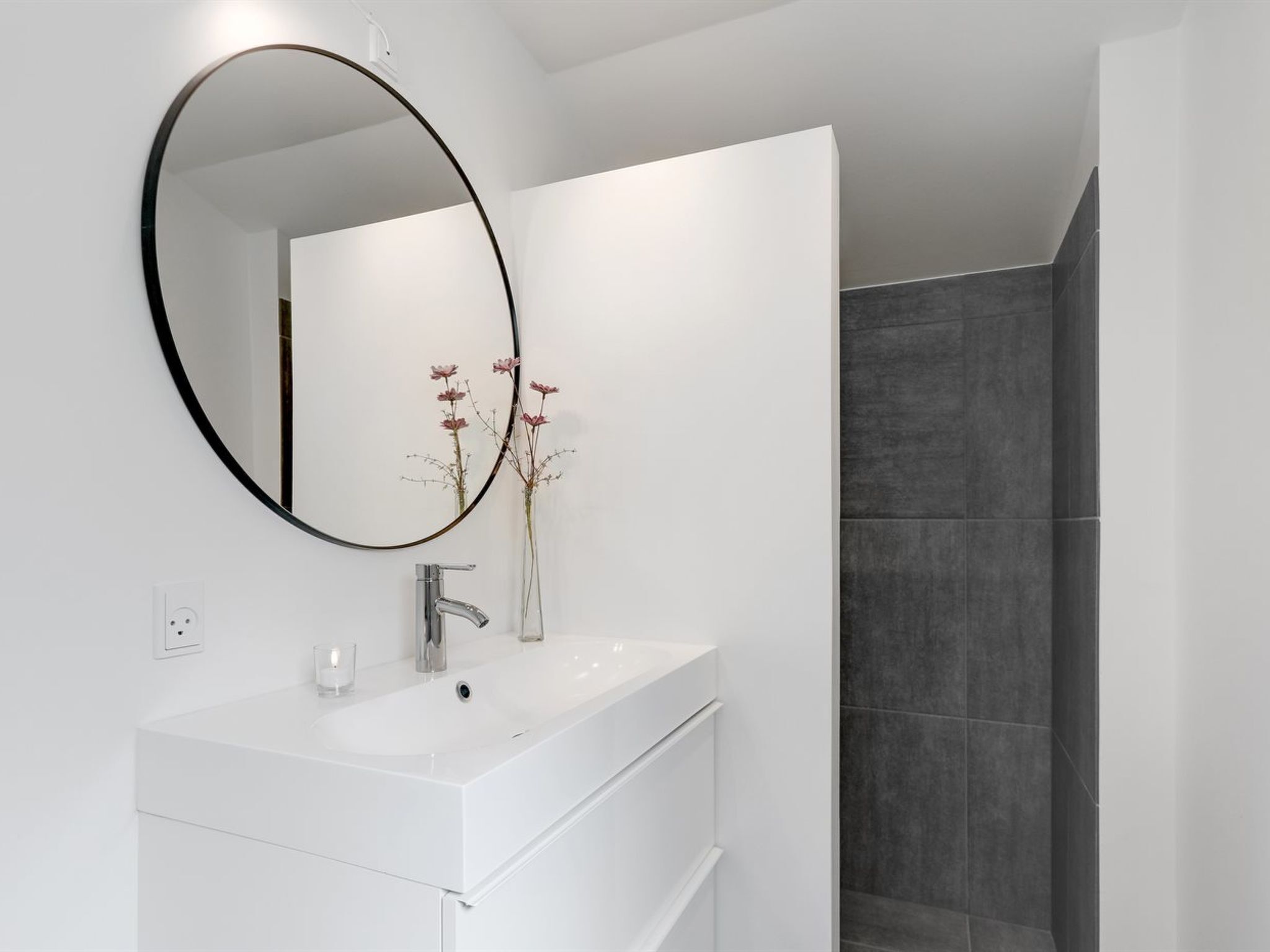 A modern bathroom with a white vanity, a round mirror, and a shower area with dark tiles.