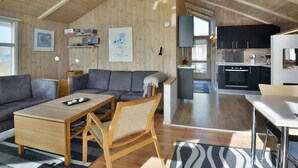 A spacious living area with a sofa, coffee table, and a dining table. A kitchen with modern appliances and black cabinets is visible in the background. - "Styrbiorn" - 100m from the sea by Interhome (Løkken)