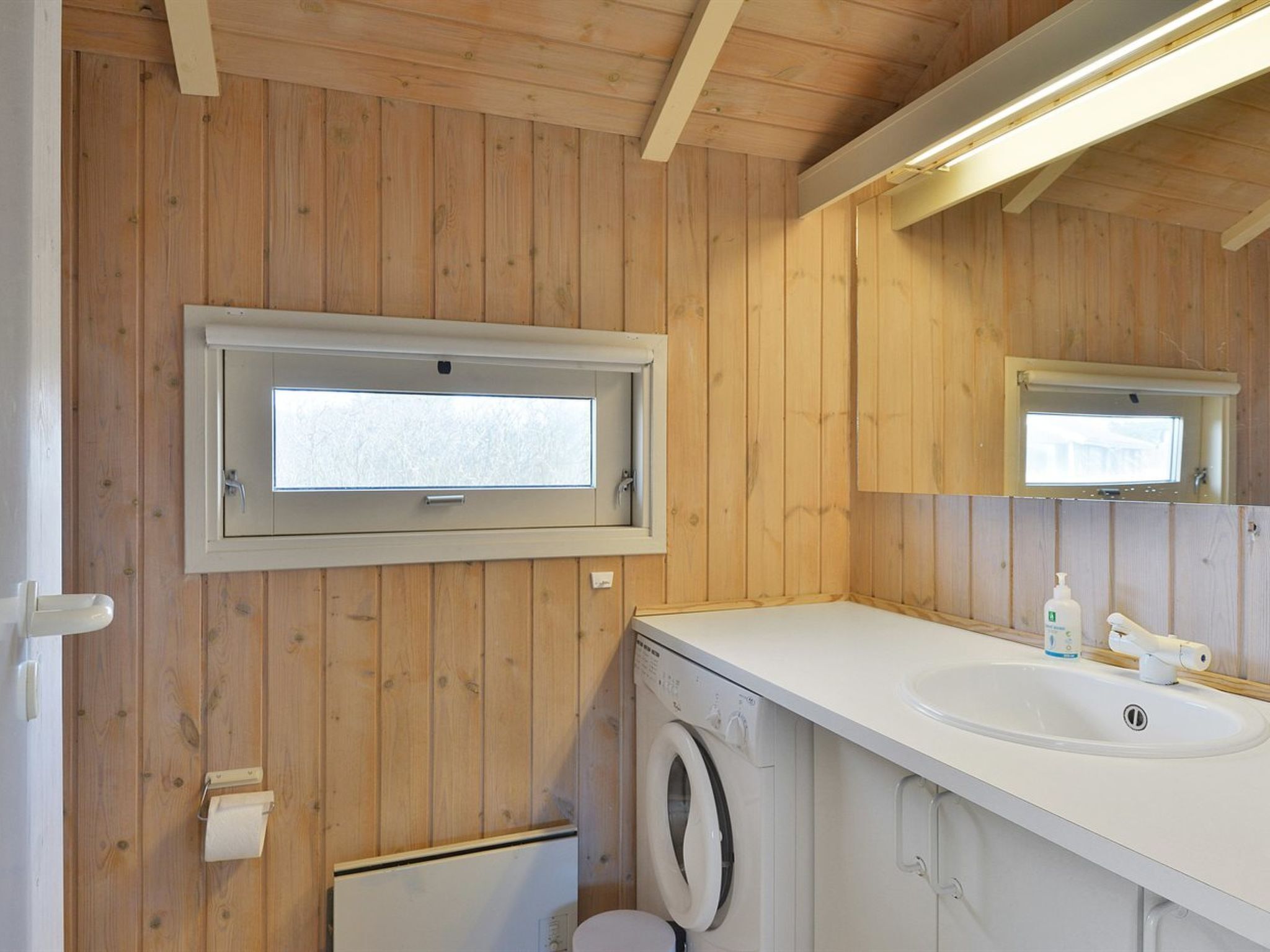 Un baño pequeño con paredes de madera, un ventanal integrado, una lavadora y un lavamanos con un espejo encima.