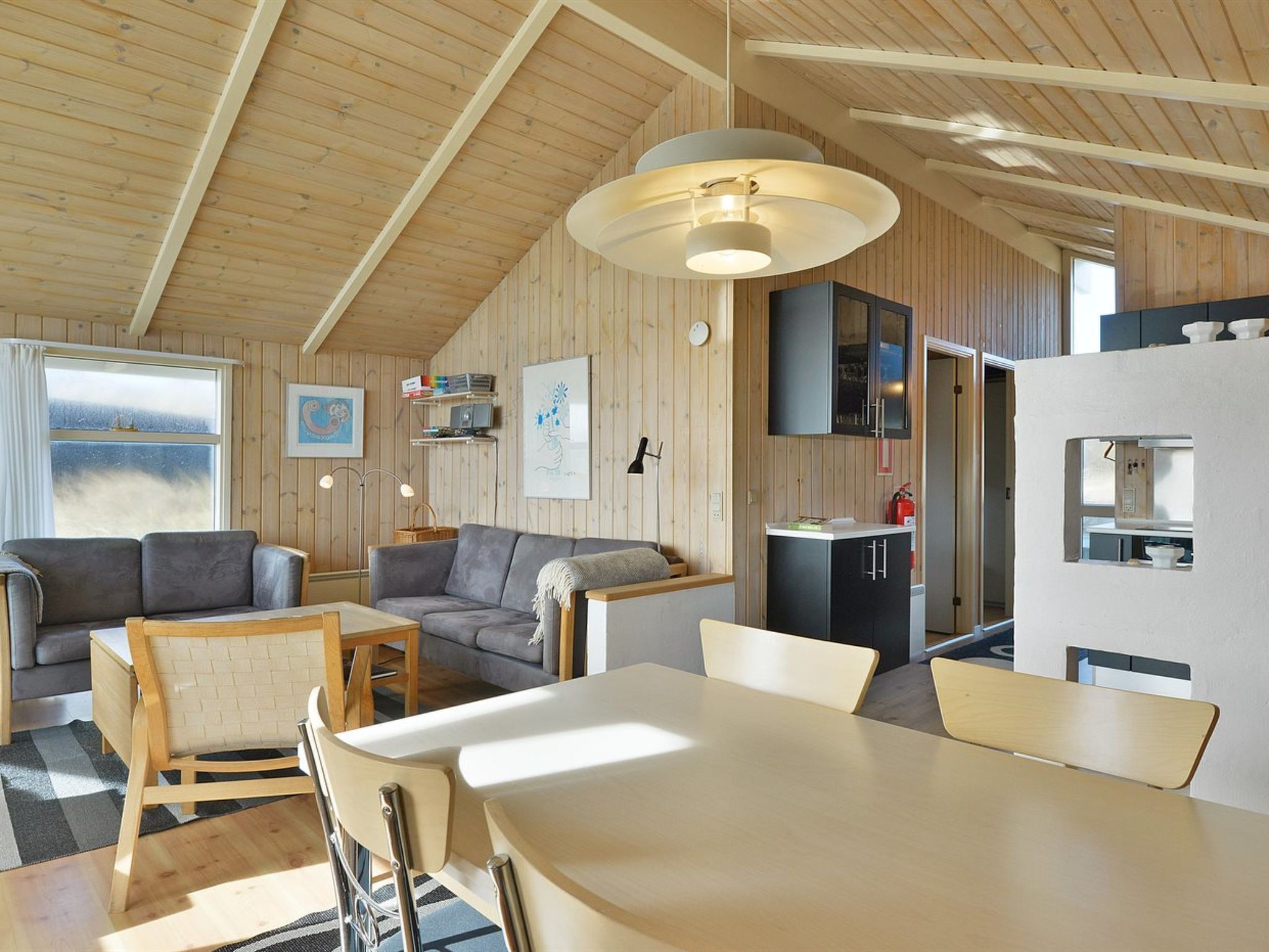 Un intérieur de chalet chaleureux et bien éclairé, comprenant un coin repas, deux canapés, une cuisine et une grande fenêtre.