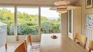 A dining area with a long wooden table, cushioned chairs, and a pendant light. Large windows offer a view of greenery and a distant landscape. - "Ludewiga" - 350m from the sea by Interhome (Glesborg)