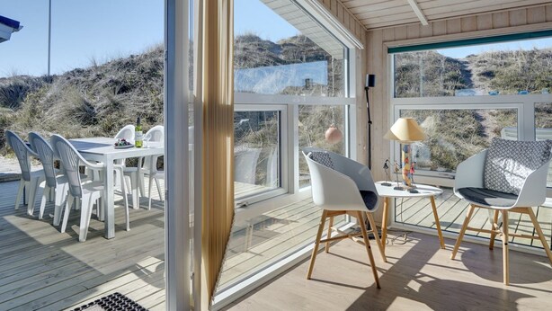 Et moderne udendørs spiseområde med glasfacade, hvide plaststole og et bord med en flaske og druer.
