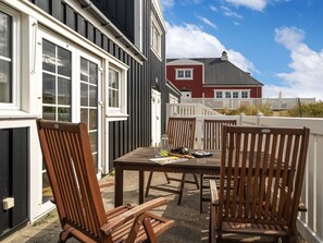 Eine Terrasse im Freien mit einem Holztisch und -stühlen, einem weißen Zaun und einem roten Gebäude im Hintergrund.