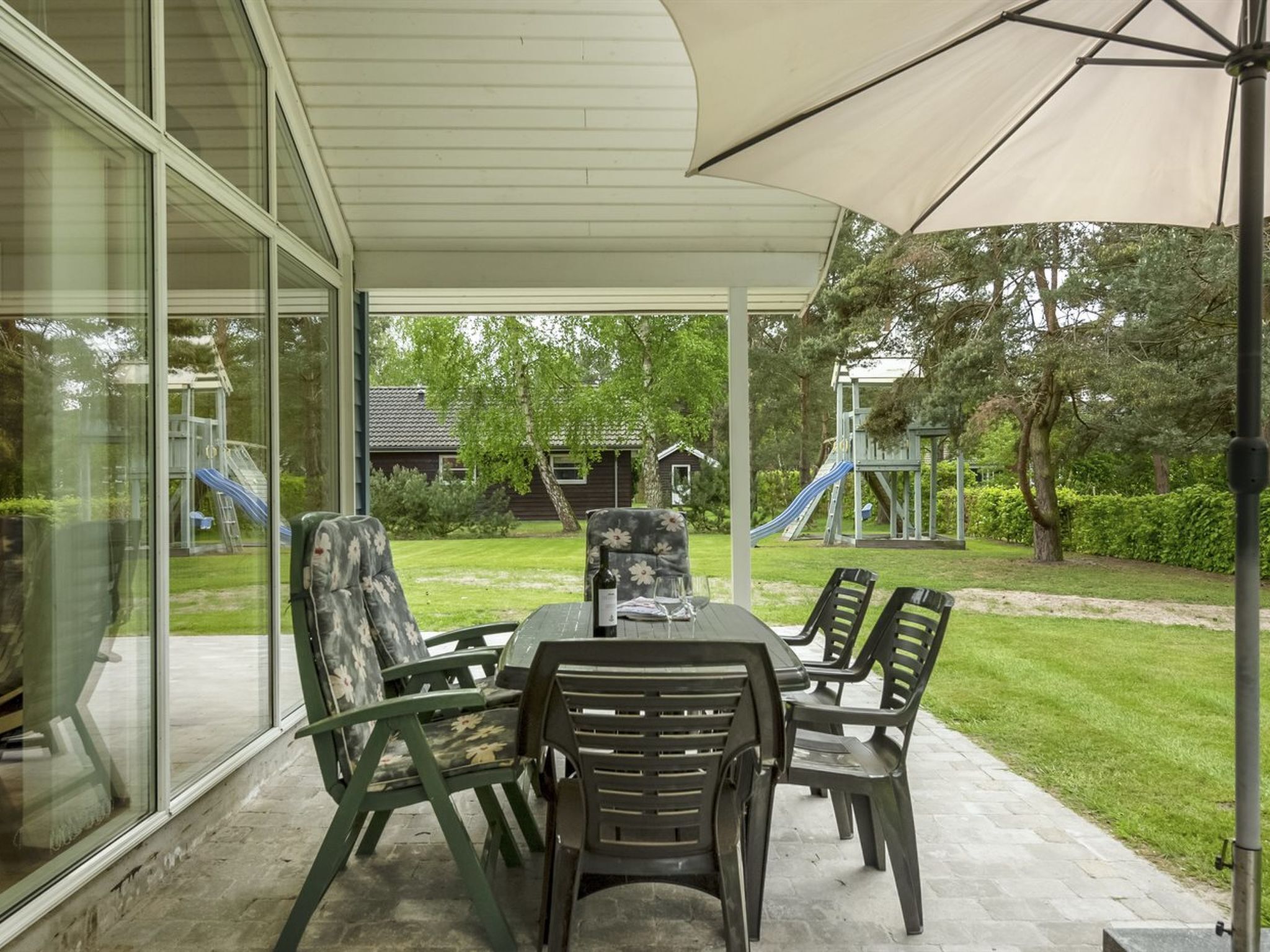 Eine überdachte Terrasse mit Tisch und Stühlen, einer Glaskonstruktion und einem Spielplatz im Hintergrund.