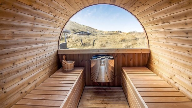 A sauna with wooden walls and benches, a view of a landscape through a semi-circular window, and a metal container on the floor.