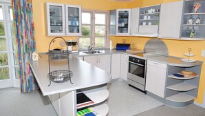 Eine moderne Küche mit weißen Schränken, einem Einbauofen und einem Spülbecken. Im unteren Schrank befinden sich Bücher und am Fenster hängt ein farbenfroher Vorhang.