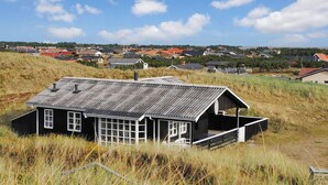 Eine rustikale Holzhütte mit geneigtem Dach, umgeben von hohem Gras und einem entfernten Blick auf eine Wohngegend mit Häusern.