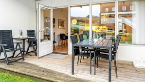 Un área de comedor al aire libre con terraza de madera, sillas negras y una puerta de cristal que da a un comedor.