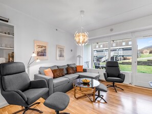 Een moderne woonkamer met een grijze bank, twee fauteuils, een salontafel met fruit, en een groot raam met uitzicht op een gebouw en groen.