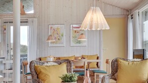 Une pièce chaleureuse, meublée de meubles en osier, avec une table sur laquelle se trouvent une théière et une bouteille. Les murs sont ornés de tableaux encadrés et une lampe est suspendue au plafond.