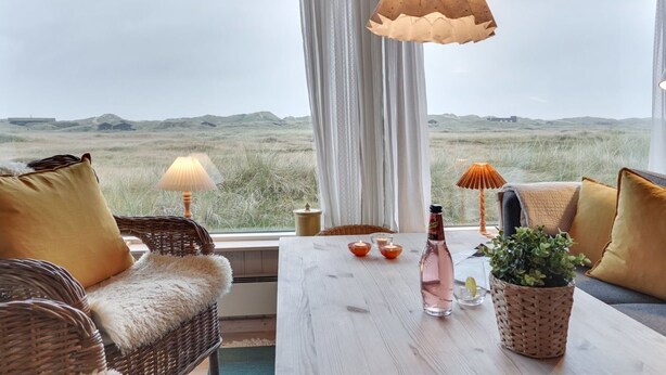 Une pièce offrant une vue sur un paysage verdoyant, avec un canapé en osier, une table en bois sur laquelle sont posés une bouteille et des bougies, et une lampe suspendue.