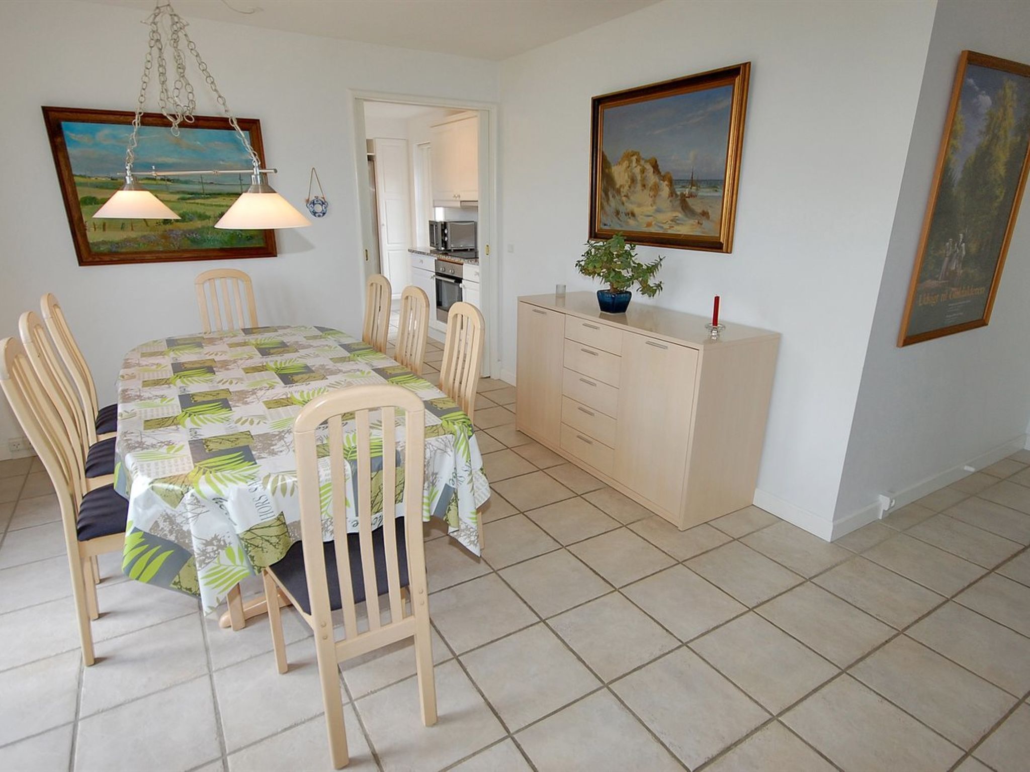 A dining area with a table set for four, wooden chairs, a cabinet with drawers, and two paintings on the walls.