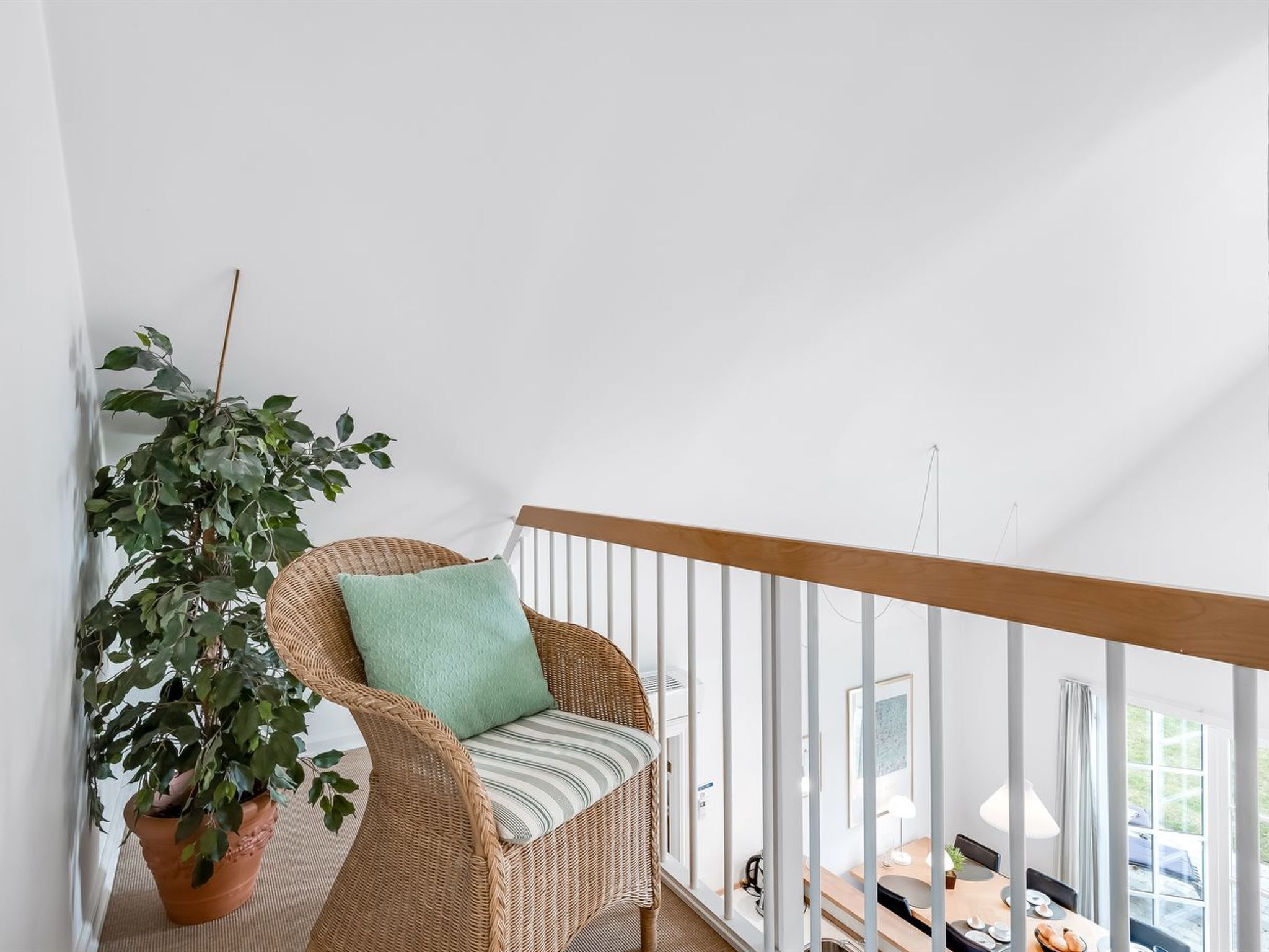 Rottinkituoli, jossa on tyyny, valkoisella kaiteella varustetulla parvekkeella. Parvekkeella on myös ruukkukasvi, ja sieltä on näkymä sisätilojen ruokailutilaan.