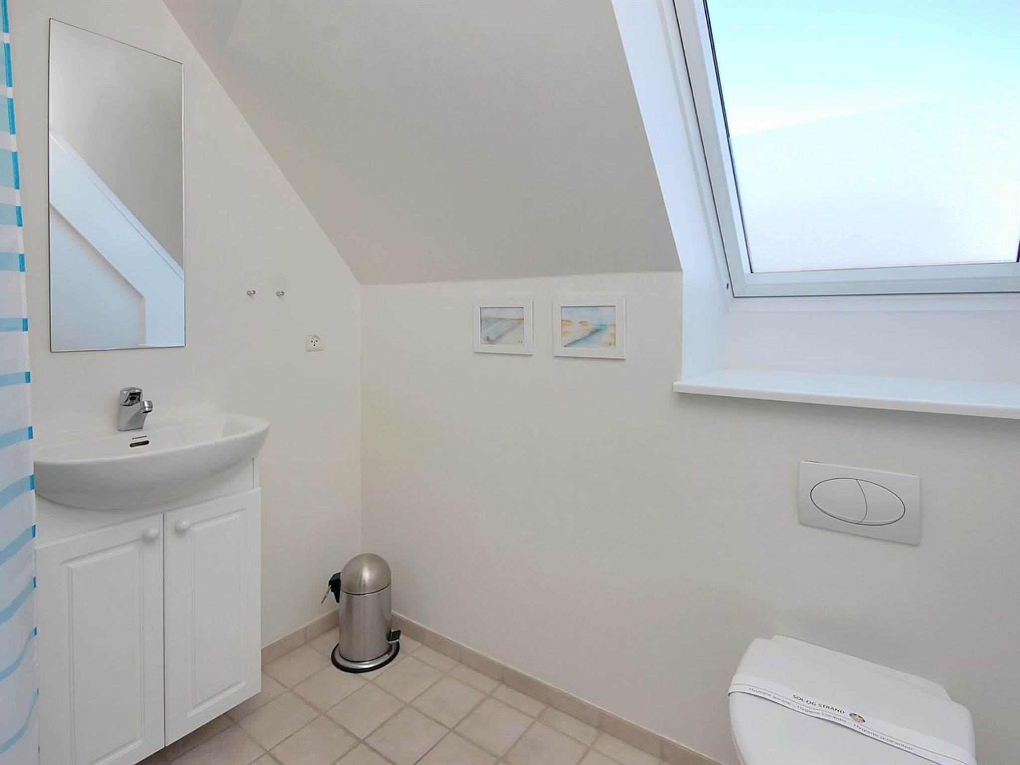 Une salle de bain moderne avec un lavabo blanc, un miroir et des toilettes. On y trouve deux tableaux encadrés au mur, une fenêtre et une poubelle.