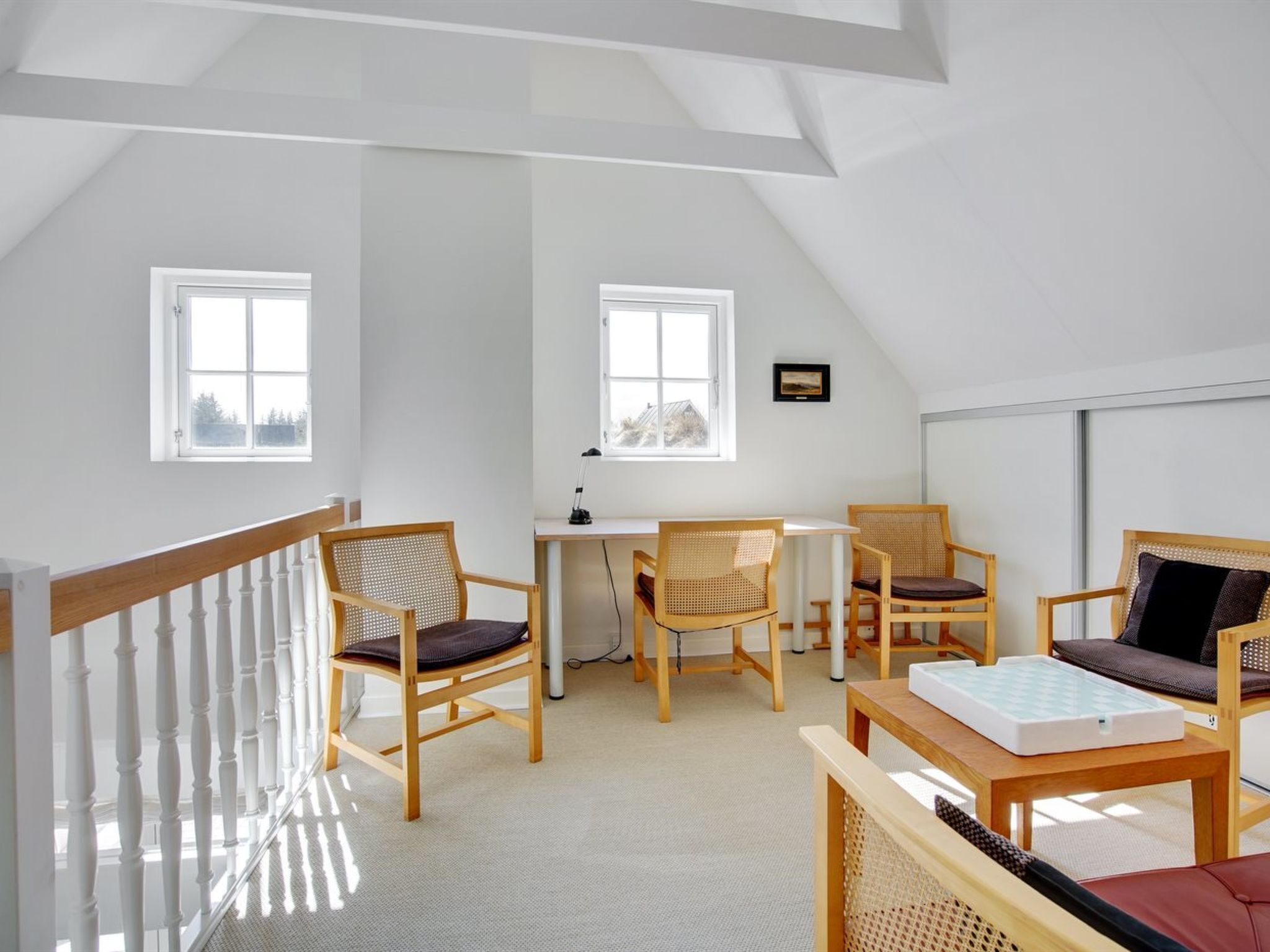 A spacious loft bedroom with a sloped ceiling, two large windows, a desk, and a cozy seating area with chairs and a sofa.