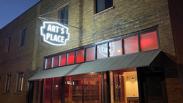 Un edifici amb un rètol que diu "ART'S PLACE" i una façana amb grans finestres i una tenda metàl·lica.