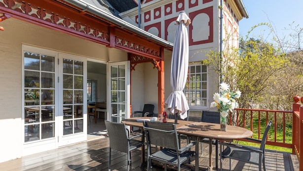 En udendørs terrasse med bord og stole, en markise og udsigt til en bygning med store vinduer og en balkon.