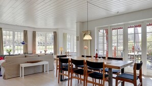 En rummelig stue med spisebord, stole, sofa og et stort vindue med gardiner.