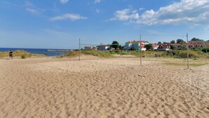 En strand med volleyballnet, sandet område og huse i baggrunden.