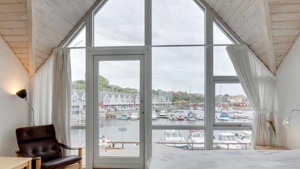 Quarto com uma grande janela com vista para uma marina, teto de madeira e cama com colcha branca.