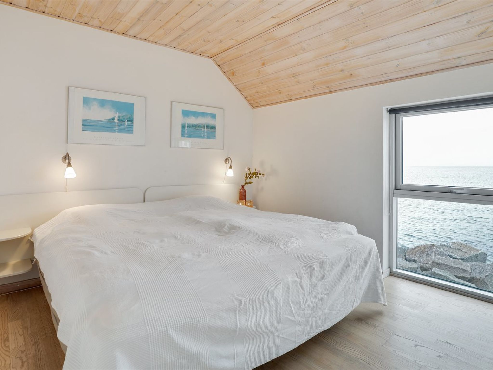 A bedroom with a large bed, wooden ceiling, two framed pictures, bedside lamps, and a window with a view of the sea.