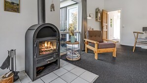 Ein Wohnzimmer mit einem schwarzen Kaminofen, einem gestreiften Sessel und Blick ins Freie durch ein großes Fenster.