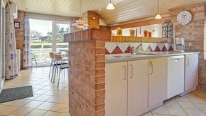 Una cocina con pared de ladrillo, armarios blancos y piso de baldosas. Hay un reloj en la pared y una ventana con cortinas.