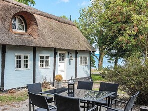 Une petite maison au toit de chaume, dotée d’un petit porche et d’une table avec des chaises devant.
