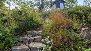 A rustic stone staircase leading to a traditional wooden building with a triangular roof, surrounded by lush greenery and wildflowers.