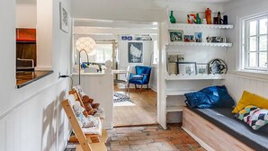 A cozy living room with a wooden bench, a blue armchair, and a colorful cushion. There is a white shelf with decorative items and a window with a view.